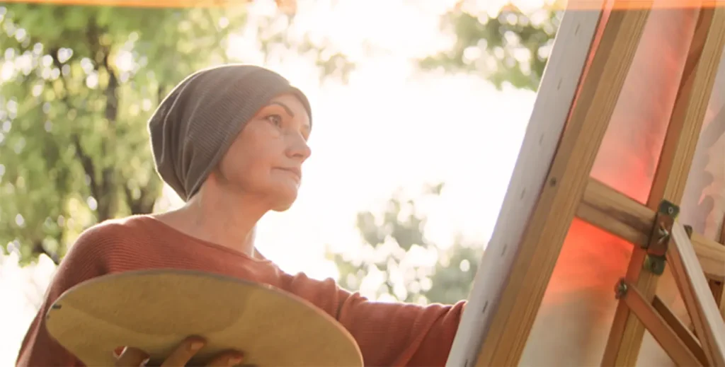 A woman painting outdoors in natural light, representing creative and expressive art as part of holistic healing at a cancer retreat.