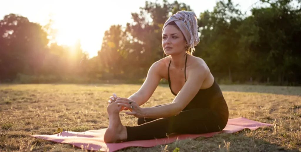 A woman practicing gentle yoga stretches on a mat in an open field, reflecting yoga and meditation for cancer healing and relaxation.